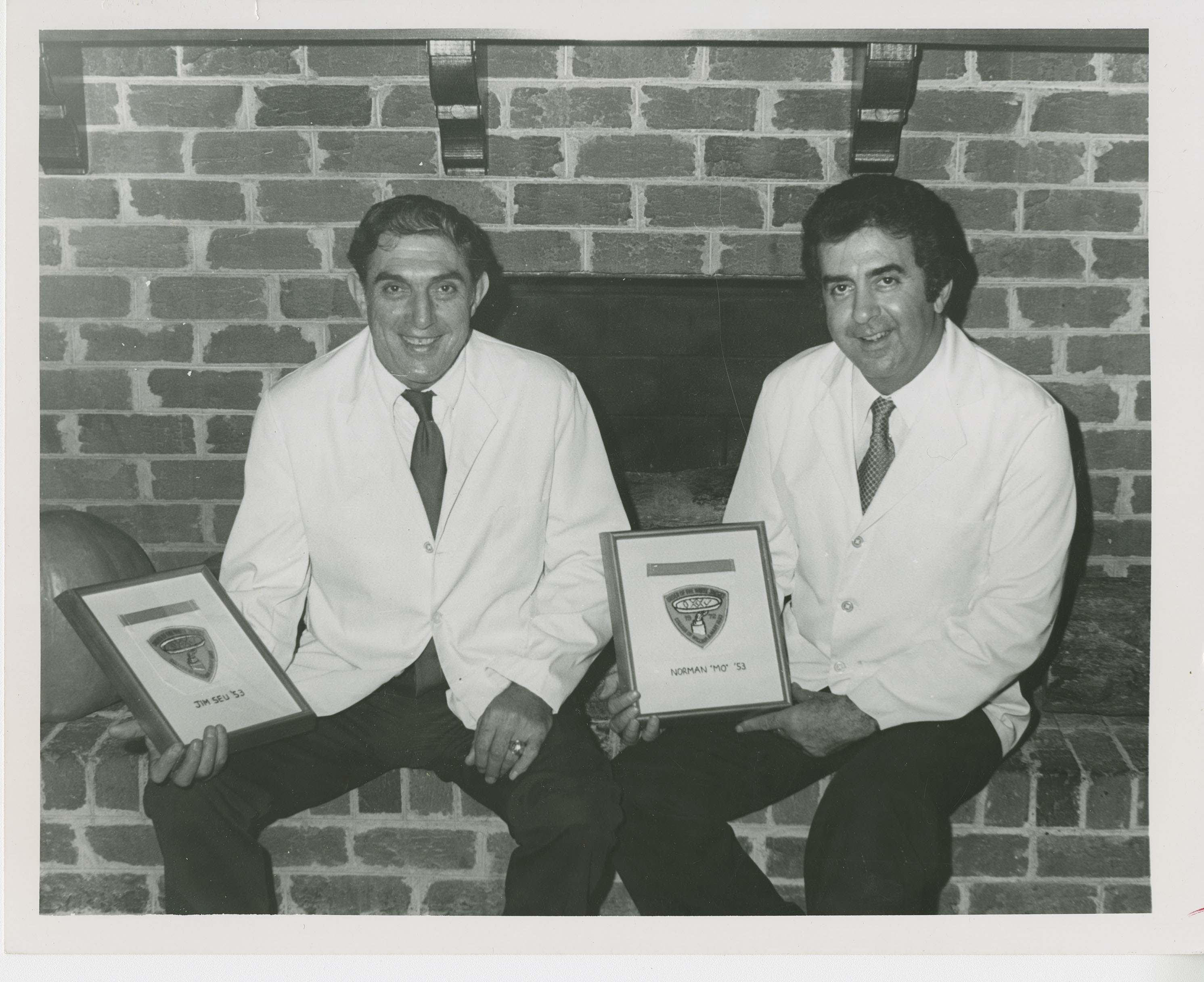 Photograph of Jim Seu and Norman Moomjian seated in front of a brick fireplace, each holding an award